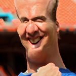 A figure stood before the vibrant haze, bearing a stretched forehead and chin. The visage called to mind memories of Peyton Manning on the field. Clad in blue, tinged with white and orange patches, he smiled. It was a grin of quiet endurance against the blurred chaos behind the football champion.