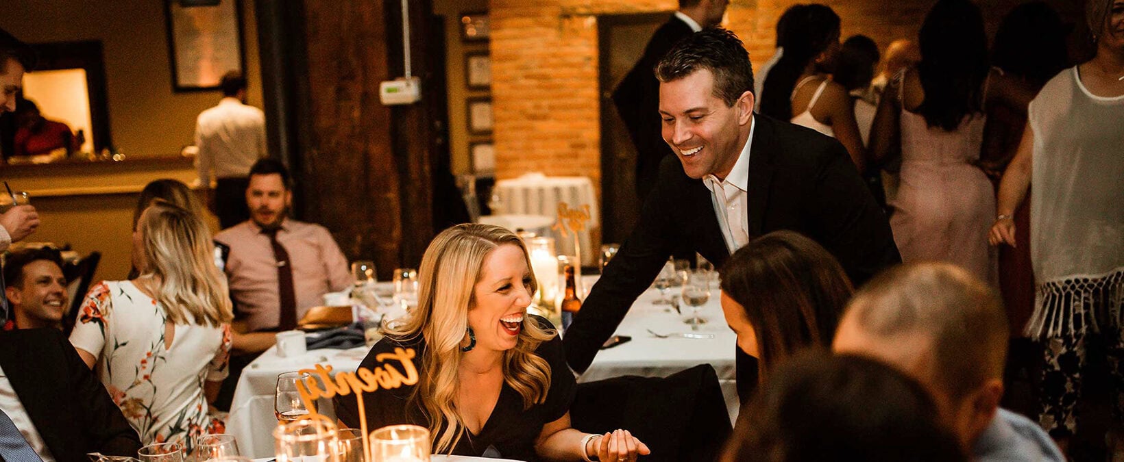 A lively dinner event in a large, rustic dining hall in Louisville. At the center of the image, a woman wearing a black dress is seated at a round table, laughing with her friends and colleagues. She and her peers appear to be entertained by the handsome close up magician in Louisville stood beside her. The entertainer is dressed in a black suit with a white dress shirt. The magician and mentalist smiles as he performs what appears to be close-up magic or entertainment for the guests at the table in Louisville.  The table is elegantly set with white tablecloths and lit candles, creating a warm, intimate atmosphere. There's a small decorative sign in the center of the table. Wine glasses and water glasses are arranged on the table in a Louisville style.  In the background, other guests can be seen seated at nearby tables, and some in the audience are standing or moving around the Louisville venue. The ambiance suggests this is a special celebration, a wedding reception, anniversary party, or upscale social gathering in Louisville. The lighting is warm and golden, highlighting the joyful expressions of the audience and guests, creating a festive, elegant mood throughout the Louisville space.